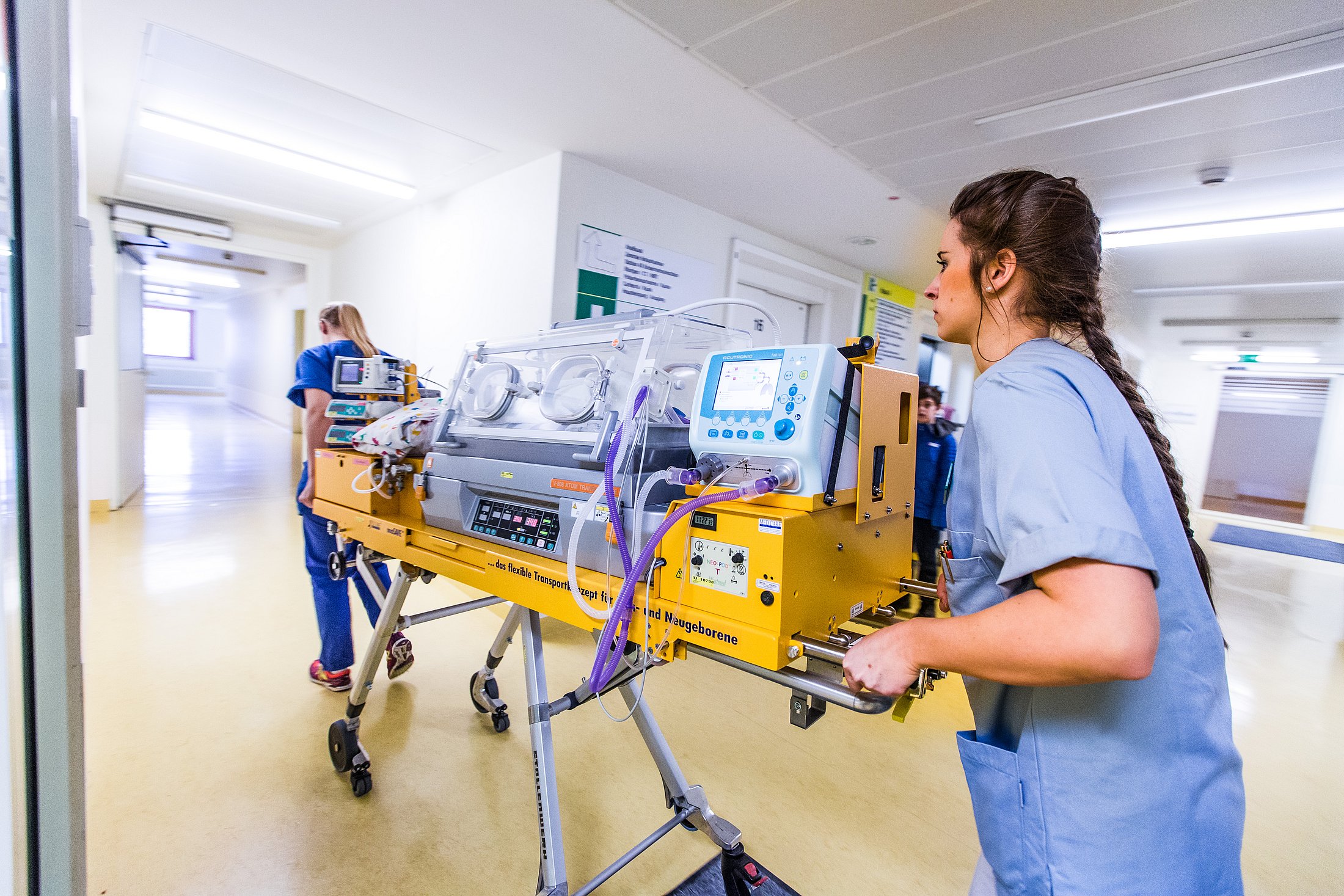 Nurse pushing a neonatal incubator in a hospital hallway.