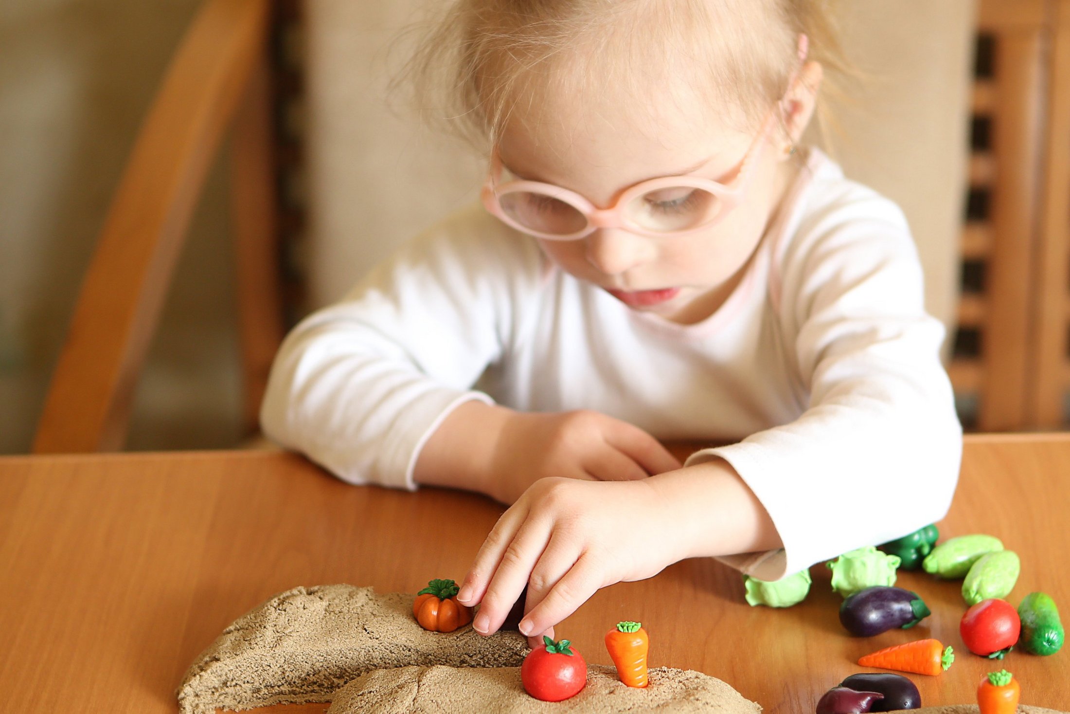 A young girl with glasses playing with toy vegetables and sand.