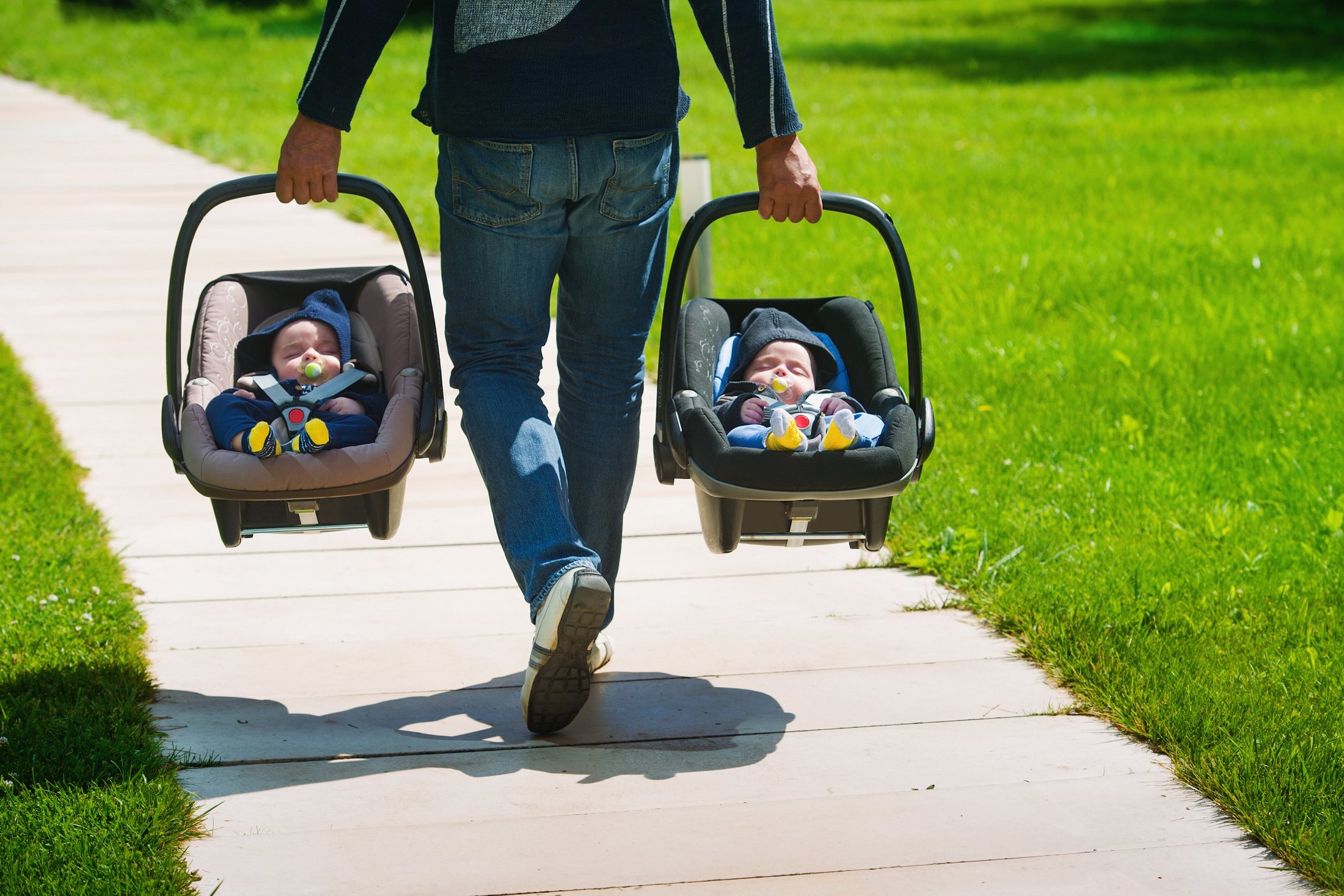 A father carrying twin babies in car seats on a park path.