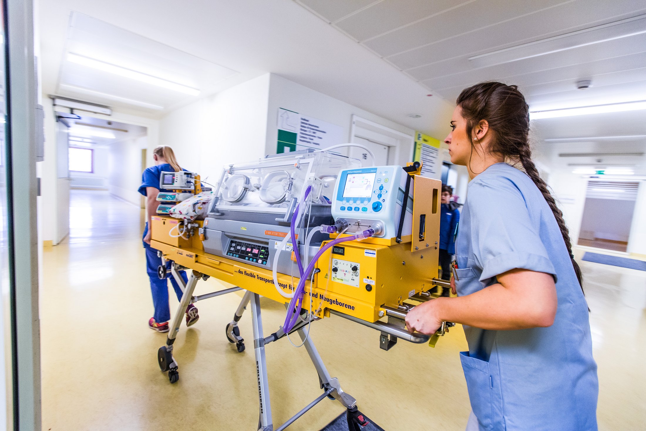 Nurse pushing a neonatal incubator in a hospital hallway.