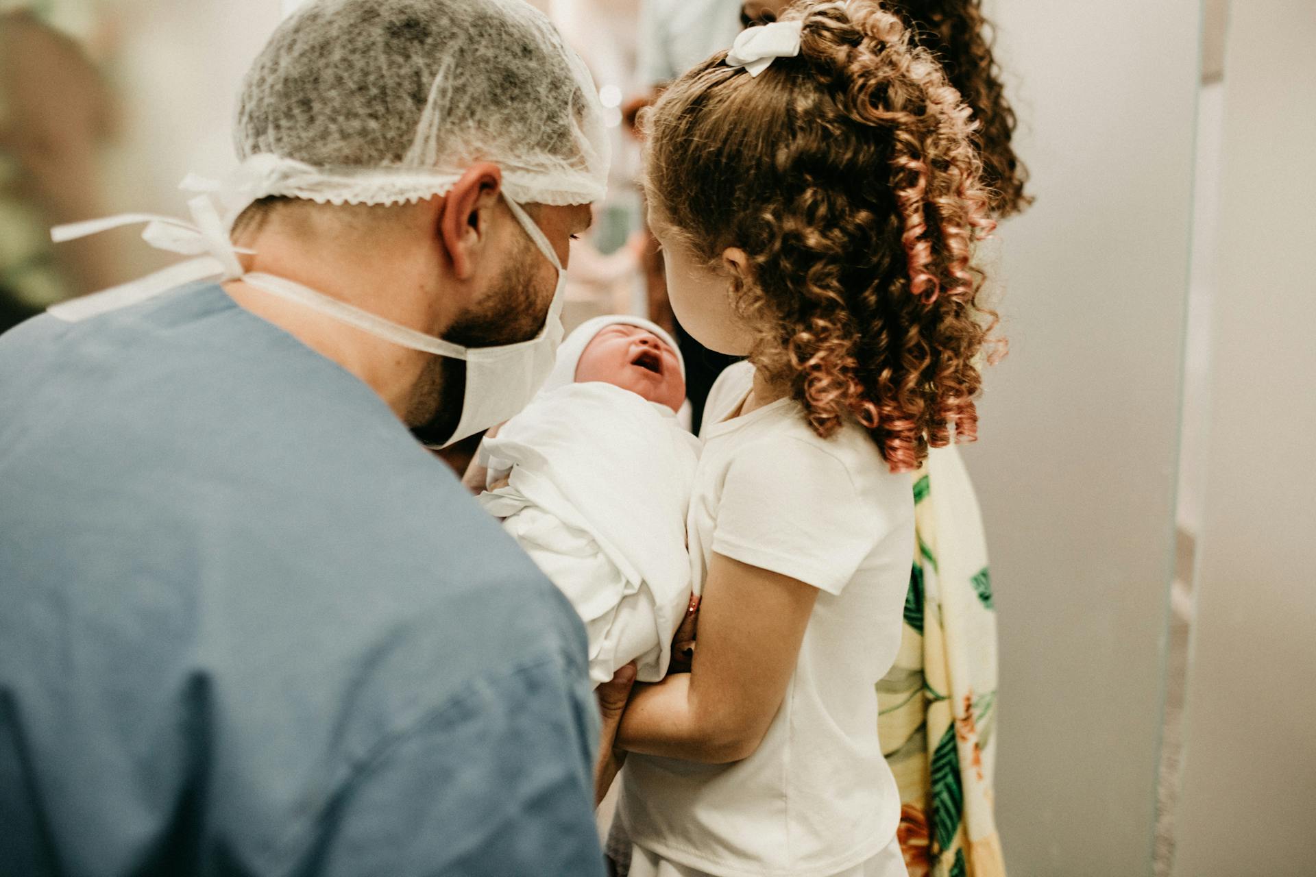 Child holding a newborn baby with a masked healthcare worker nearby