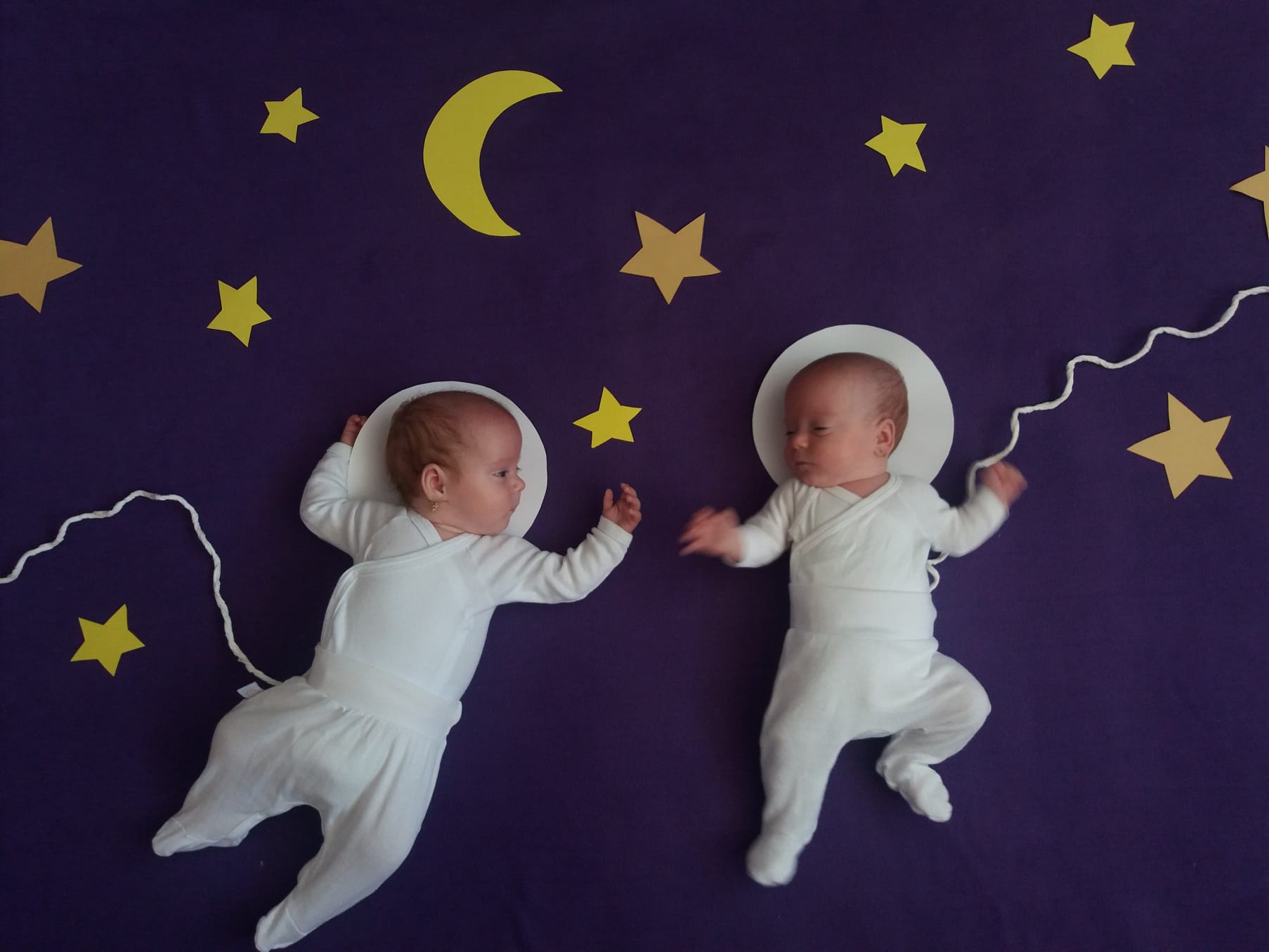 Diana and Rebeca, twin sisters born preterm, dressed in white and lying on a purple blanket decorated with stars and moons.